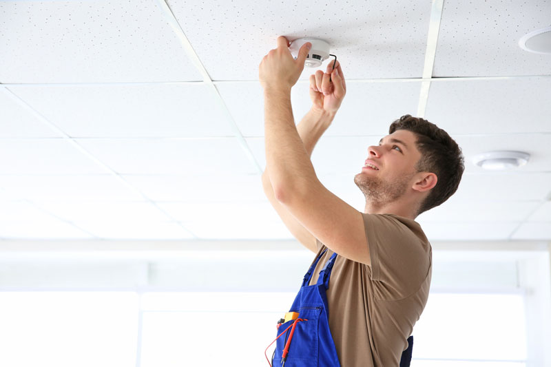 Young electrician maintaining smoke detector on ceiling