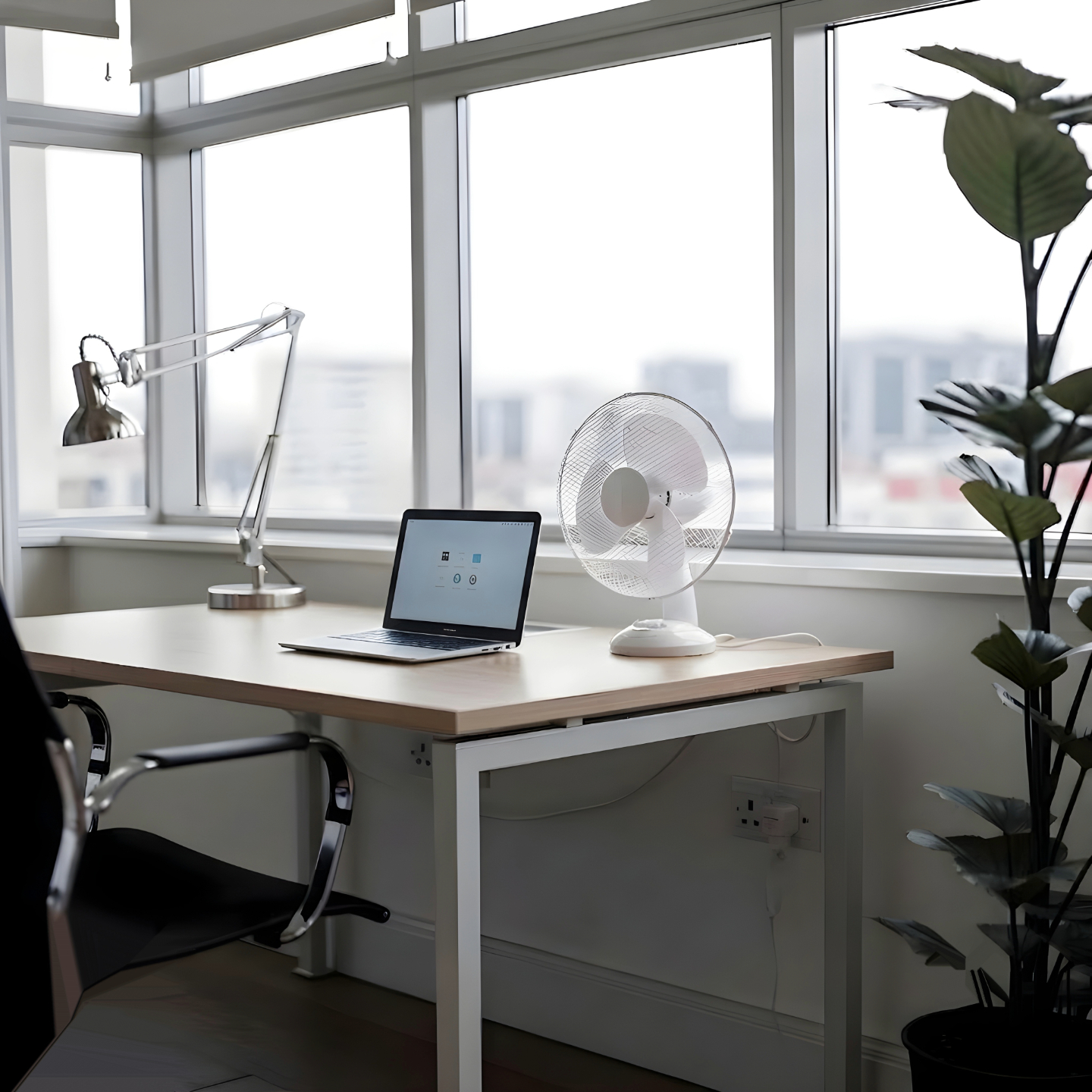 White desk fan on a home office desk next to a laptop and lamp beside a large window